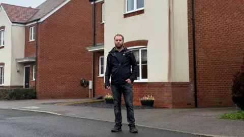 BBC Dean Carpenter standing in front of his new build home with a large dark red stain running down one corner of the walls