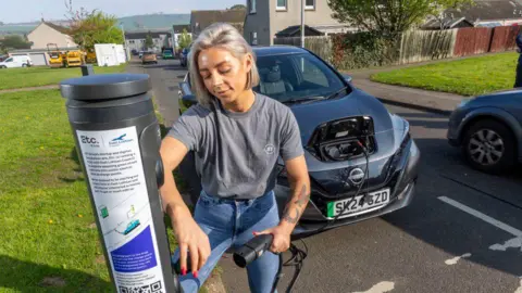 BT Group A woman plugs her electric car into the public charging point on the street in a housing estate.