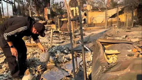 Man in black track suite leaning over and holding vase amid burned ruins 