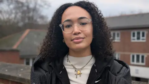 BBC Olimata stands outdoors facing the camera and has long, curly black hair and glasses, wearing a black jacket, white top and jewellery around her neck