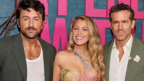 Getty Images Three people - Brandon Sklenar, Blake Lively and Ryan Reynolds - are dressed in formal attire and posing for a photo. They are stood in front of a large sign that reads 