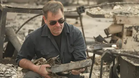 BBC Peter Mitchell has a short beard and brown hair, with a dark shirt. He sits looking at the remnants of a piece of furniture - surrounded by the burnt out shell of his home