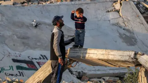 Getty Images A man speaks with a boy standing on a broken concrete beam by the rubble of a collapsed building at a camp for people displaced by conflict in Bureij in the central Gaza.