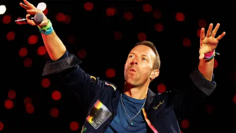 Getty Images Chris Martin from Coldplay holding both hands up in the air, with a black microphone in his right hand. he is wearing a denim jacket and blue top underneath, with red spotlights behind him.