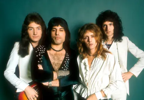 Rock band Queen pose backstage at the BBC in the 1970s.. Singer Freddie Mercury has his arms folded, while wearing a black catsuit. The other members are dressed in white.