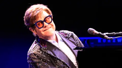 Getty Images Elton John plays piano while dressed in a purple sequinned outfit