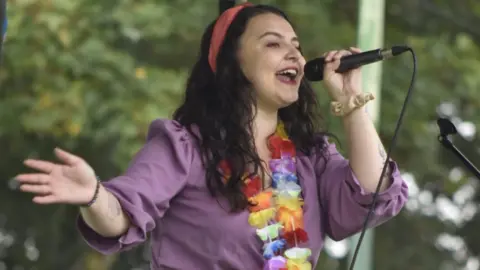 Kelley Tigerheart A woman with long black hair and wearing a pink head band, a purple shirt and flowers around her neck, sings into a microphone on stage.