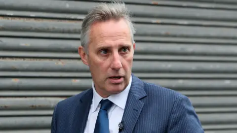 PA Media Ian Paisley - a man with short grey hair wearing a striped blue suit, a blue tie and a white collared shirt is standing in front of grey metal shutters.