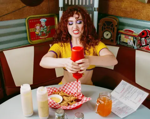 Ryan Lee Clemens Chappell Roan, on the set of the video for Hot To Go, squeezes ketchup onto a basket of fried chicken, in a typical American diner