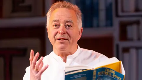 PA Media Frank Cottrell-Boyce on stage, gesturing with one hand and holding one of his books in the other