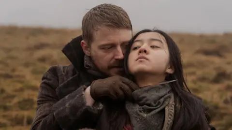 Lionsgate UK/HanWay Films Still from upcoming film Tornado showing Little Sugar (Jack Lowden) and Tornado (Kōki) - Little Sugar holds a blade at Tornados neck as he stands behind her on a grass covered hill with grey skies behind them.