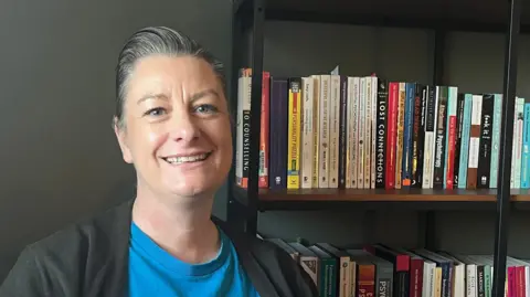 BBC Zoe Hughes, an Exeter City council member, wearing a blue T-shirt and a black top, sitting next to a bookshelf