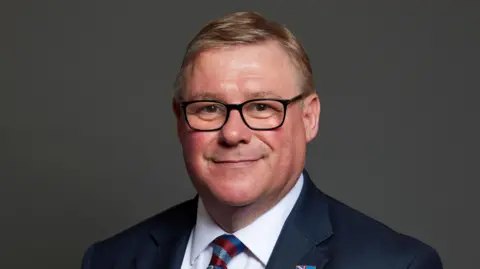 House of Commons/Mark Francois Mark Francois MP's official picture portrait from the House of Commons. He has short brown hair and black glasses and is smiling. He is wearing a blue jacket, a blue shirt and a patterned tie.