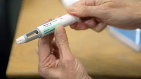 A pair of hands holding a Wegovy injection pen, which consists of a white plastic body with a dial and a window and a needle at the end. The label reads 