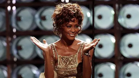 Getty Images Whitney Houston performing on stage during the 2004 World Music Awards at the Thomas and Mack Center, Las Vegas in 2004. She is wearing a gold strappy dress.