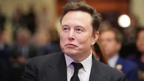 Getty Images Elon Musk, who has dark hair, is wearing a dark suit, white shirt, and a black tie in a head and shoulders shot. He is photographed looking to his right, with a crowded room of people in the background. 