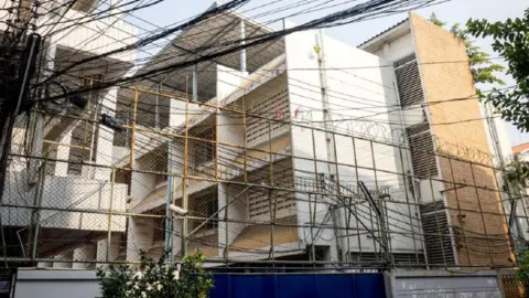 Getty Images A tall meshed fence topped with barbed wire guards the immigration detention centre where 48 Uyghurs are being held in Bangkok - the building is behind a blue gate and has walls that are white, alongside some built with brown bricks. 