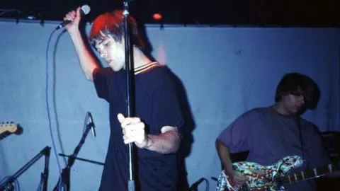 The New Adelphi Club The Stone Roses during a live performance. Frontman Ian Brown, on the left, is wearing a navy t-shirt and is holding a mic up to the ceiling with his right hand and a mic stand with his left. On the right is bassist Mani. He is wearing a blue shirt and playing on a bass guitar.