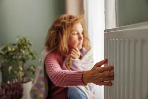 Getty Images Woman wearing a jumper and wrapped in a blanket puts her hand on a radiator. A plant is in the background.