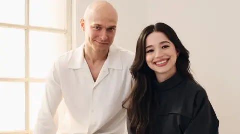 Getty Images Yura Borisov and Mikey Madison of Anora at the Toronto International Film Festival. They are standing in front of a window. Borisov is wearing a white shirt, while Madison is dressed in black.