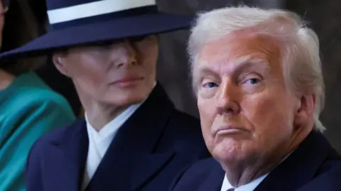 Reuters US President Donald Trump looks down the lens on the inauguration day of his second Presidential term with first lady Melania behind him, wearing a large navy hat.