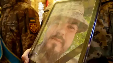 BBC/Xavier Vanpevenaege A soldier holds a framed photo showing Andriy Kusmenko smiling at the camera wearing a camouflage hat