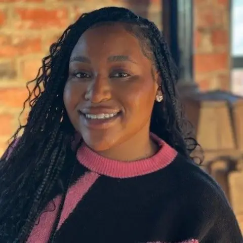 Singer Sisi is wearing a navy blue jumper with pink collar and design and has long dark braided hair. She is smiling, standing front of a brick internal wall which is blurred out.