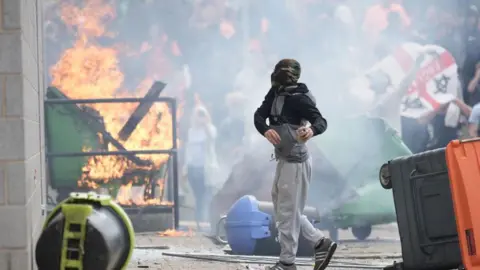 Getty Images A masked man holds something in his hands as a building is set on fire