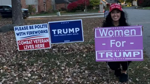 Amanda Sue Mathis Amanda Sue Mathis wears a red Make America Great Again hat while crouching behind a purple Women for Trump sign in her yard, which is full of autumn leaves. Other signs include a navy Trump campaign sign and a sign that says: 