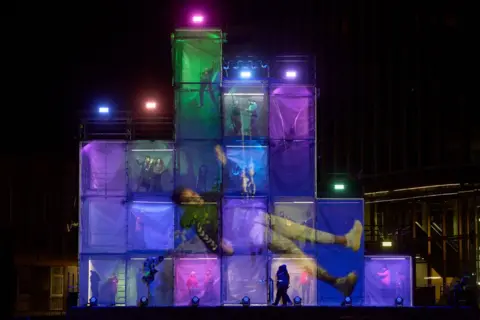 David Levene/Bradford City of Culture A shot of the stage showing towers made up of a series of colourful boxes with performers in them and a projection of a large figure across them all on the front