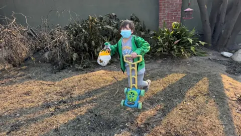 A young boy plays with a toy lawnmower wearing a mask