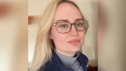 Sophina A woman with long blonde hair wearing glasses is looking to the side of a camera. She is wearing a blue top over a grey roll neck top.