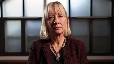 A portrait of lawyer Sally Marsden. She is sitting in front of an arched window and has blonde hair. She is wearing a statement necklace, black top and maroon jacket.