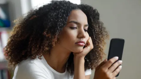 Getty Images A young woman with shoulder-length hair wearing a white t-shirt, staring at her phone while looking fed up.