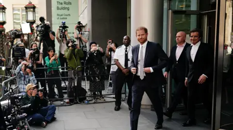 PA Media Prince Harry leaves the court during his hacking case against the Daily Mirror. He wears a dark coloured suit, white shirt and tie. His barrister David Sherborne, also dressed in a dark suit is on his left.  A crowd of photographers are behind a metal barrier