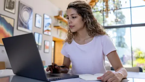 Getty Images A woman looking in to a laptop with a pen and paper nearby