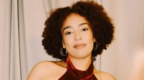 Jamie Drew @jdshotyou A woman with dark curly hair and wearing earrings and a red dress smiles as she stands in front of a curtain.