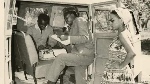 Herbert Weiss Pierre Mulele, one foot inside a van, peruses a piece of paper. A man inside the van uses a typewriter, while Andrée Blouin stands outside the van with her hands on her hips.