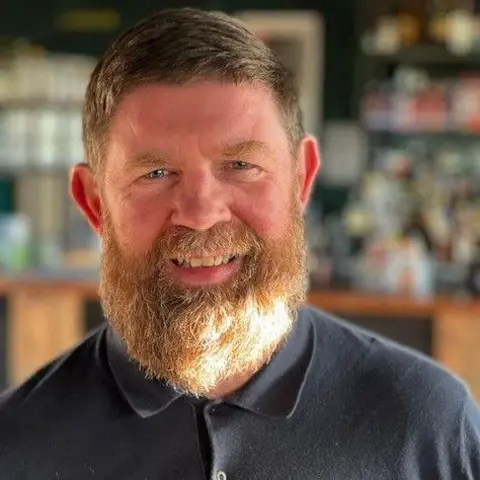 Jim Mawdsley, the principal advisor for events and culture for Newcastle City Council, smiling. He has short light brown hair and a bushy, lighter brown beard and moustache. He's wearing a dark blue polo shirt. The background is blurred.