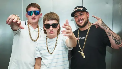 BBC/Jonathan Birch Sam Robinson (Clive), Gareth Kelly and Kane Welsh from Bad Boy Chiller Crew pose, with their fingers doing salute signs. The trio are wearing gold chains around their necks, with Clive and Kane also wearing sunglasses and pale tshirts. Gareth is wearing a black tshirt and cap.