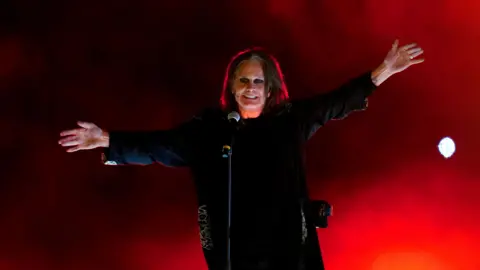 David Davies/PA Wire A man stands in front of a microphone against a red stage backdrop. He has chin-length dark hair and he is wearing a black long-sleeved tunic. He has black eyeliner around his eyes.