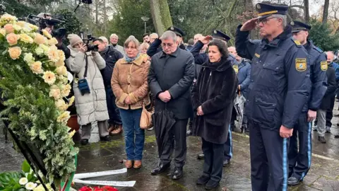 Reuters A police officer salutes and other people look on after a wreath of flowers is laid on a rainy day in a park in Bavaria where a toddler and a man were fatally attacked.