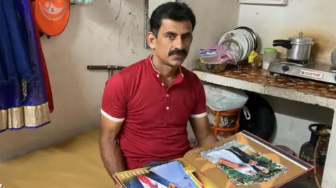 Nimisha's husband Tony Thomas wearing a red polo t-shirt and holding their wedding album is sitting on a plank placed near their kitchen. 