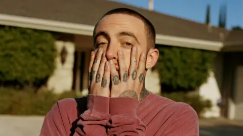 Rolling Stone/ Getty Images Mac Miller poses with his hands on his face for a magazine shoot. He's pictured outside a single-storey house on a blue-sky day. He has croppped brown hair, brown eyes and tattooed fingers and wears a red sweater. 