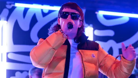 Getty Images Clive 'Sam' Robinson of Bad Boy Chiller Crew on stage, wearing an orange puffer jacket, singing into a black microphone while wearing black sunglasses. There are neon blue lights in the background.