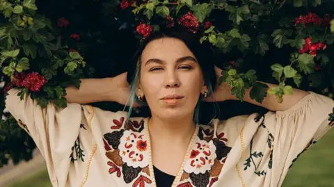 Anhelina Hlukhova Nastya Zukhvala next to a leafy bush with red flower looking directly into the camera. 