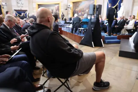 Getty Images A man in a hoodie, basketball shorts and sneakers sits in the Capitol Rotunda surrounded by men wearing suits