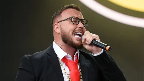 Getty Images Gareth performs at Leeds Festival in 2022. He is singing into a black microphone, wearing a black blazer, white shirt and red tie.