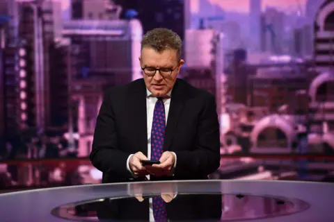 Tom Watson in a BBC studio looking down at his phone. He wears a dark suit jacket, a white shirt and a patterned tie. He has glasses on and has short hair 