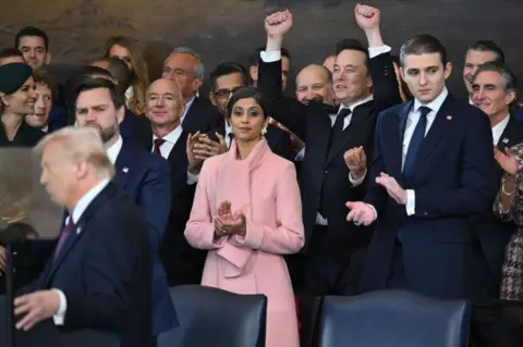 Getty Images A crowd of people in dark suits surround Donald Trump, including Elon Musk, who has his fists raised in jubilation 